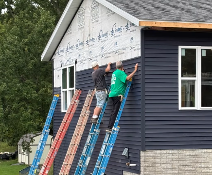 Two men on ladders leaning against a half-sided external wall of a house. One man is wearing a green shirt that says "Veridian Gives Back" on the back.